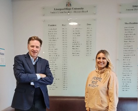 A wearing a suit and a woman wearing a hoodie stand in front of a roll of honour board.